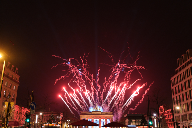 Eine belebte Straßen Szenerie in Berlin an Silvester, voller Menschen, Fahrzeuge und Gebäude, beleuchtet von Feuerwerk und Gebäudelichtern, schafft eine festliche Atmosphäre.