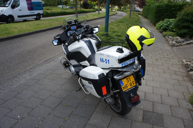 Ein polizeiliches Motorrad am Straßenrand geparkt mit einem Helm darauf, mit Bäumen, Pfählen, Gebäuden, Pflanzen, Gras, Steinen und anderen Fahrzeugen im Hintergrund.
