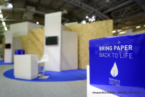 Ein Messestand mit einem blauen und weißen Schild, auf dem 'Bring Paper Back to Life' steht, auf der rechten Seite und im Hintergrund leicht unscharfe Stühle, Tische, ein Fernseher und Deckenleuchten.