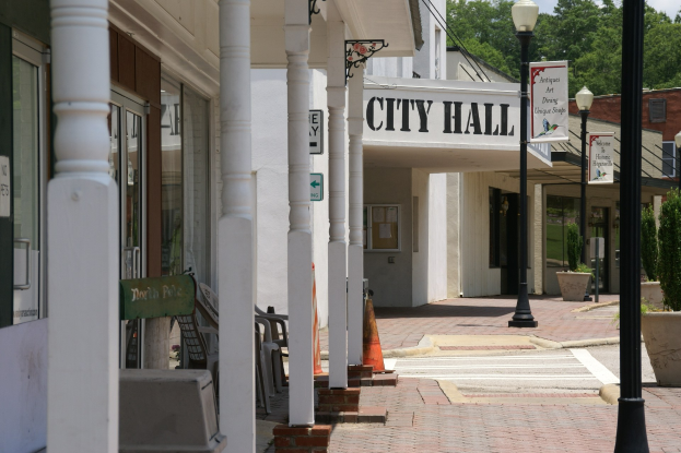 Rathaus mit umliegenden Gebäuden, Straßeninfrastruktur einschließlich Masten, Laternen und Namensschildern, Kraftfahrzeuge, Verkehrskegel, verstreute Stühle, Zimmerpflanzen, Bäume und ein bewölkter Himmel.