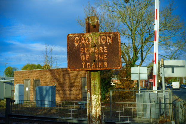Vorsichtsschild auf einer Bahnschiene, umgeben von einem Metallzaun, Bäumen, Strommasten, einem Gebäude mit Fenstern, Containern, Fahrzeugen auf einer Straße und einem bewölkten Himmel.