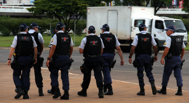 Eine Gruppe von Beamten in Uniform, einschließlich Mützen, Hemden, Schutzwesten, Uhren, Hosen und Schuhen, die auf einer Straße mit einem geparkten Lastwagen auf der rechten Seite neben einem Zaun, Gebäuden, Bäumen, Pflanzen, Pfählen und Schildern im Hintergrund und einer Brücke im oberen rechten Eck gehen.