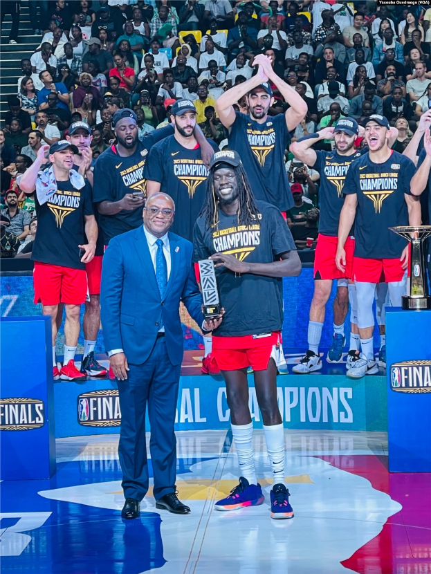 Eine Gruppe von Männern auf einem Basketballfeld, zwei mit Trophäen, mit Zuschauern im Hintergrund und zwei Trophäen auf Podesten rechts.