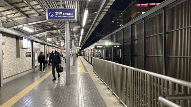 Ein Zug, der auf einem Bahnsteig einfährt, mit Menschen in der Nähe, einem Geländer auf der rechten Seite, Gebäuden im Hintergrund, Bahnsteiglampen und einer Abfahrtsanzeige.