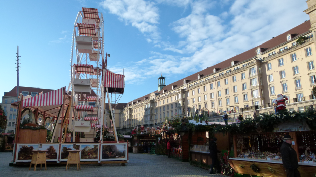 Ein lebendiger Weihnachtsmarkt in einem Stadtplatz, mit einem zentralen Riesenrad, umgeben von Gebäuden, Bäumen und einem bewölkten Himmel, mit zahlreichen Menschen und verschiedenen Gegenständen, die über das Gebiet verstreut sind.