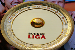 Ein Mann hält ein goldenes Schild mit «Bundesliga» darauf, mit einem Banner im Hintergrund mit drei Personen.