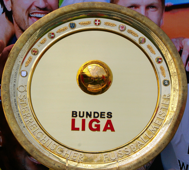 Ein Mann hält ein goldenes Schild mit «Bundesliga» darauf, mit einem Banner im Hintergrund mit drei Personen.
