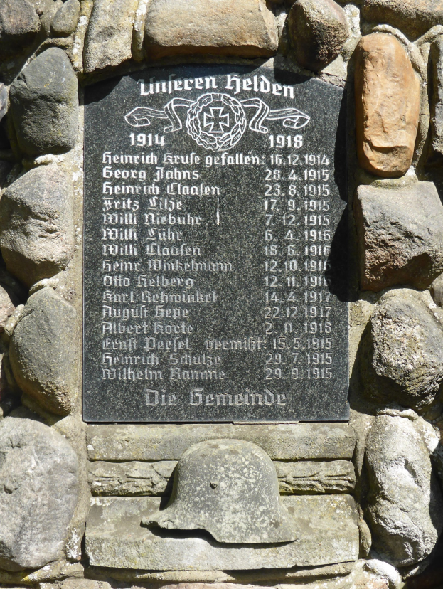 Steinwand mit einer Inschrifttafel und einem Helm darauf, wahrscheinlich ein Denkmal für deutsche Militärangehörige aus dem Zweiten Weltkrieg.