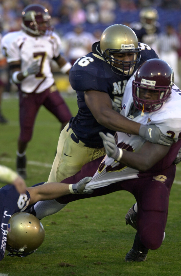 Zwei Fußballer in Helmen und Uniformen kämpfen auf dem Feld, während andere zuschauen.