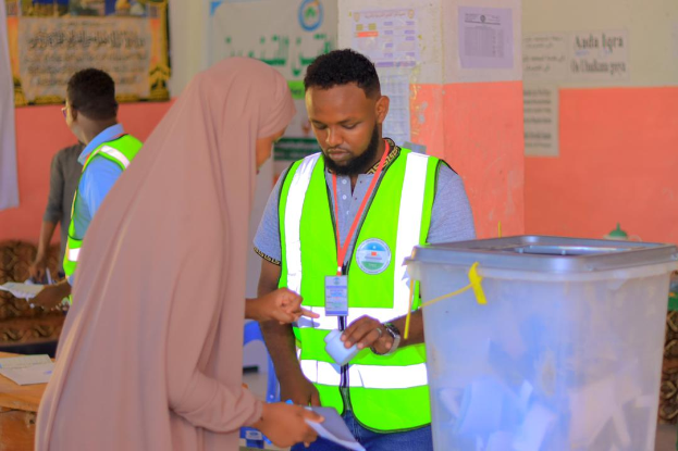Ein Mann in einer grünen Weste und eine Frau in einem pinken Kleid stehen an einem Abstimmungstisch mit Papieren und anderen Gegenständen, mit einer Wand aus angeklebten Papieren und einem Müllcontainer im Hintergrund, während der Mann ein Papier hält, das darauf hinweist, dass er abstimmt.