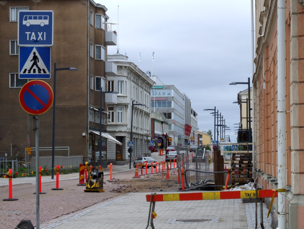 Stadtstraße mit Gebäuden, Straßeninfrastruktur, Fahrzeugen, einer Baustelle, Bäumen und bewölktem Himmel.