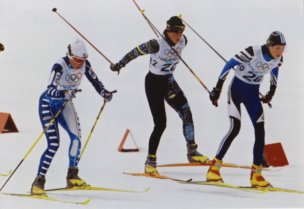 Drei Skifahrer sausen eine schneebedeckte Piste hinunter, jeder hält Ski-Stöcke in den Händen und trägt bunte Ski-Anzüge und Helme, während der Schnee in der Sonne glitzert.
