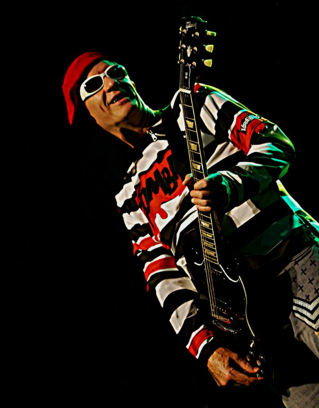 Ein Mann steht mit einer Gitarre in der Hand, trägt ein T-Shirt, eine Mütze, eine Schutzbrille und Hosen, vor einem dunklen Hintergrund.