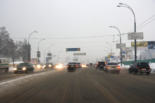 Eine städtische Straße mit viel Verkehr an einem verschneiten Tag, mit Fahrzeugen, schneebedeckter Straße, Laternen, Texttafeln, Bäumen, Gebäuden und einem Himmel im Hintergrund.