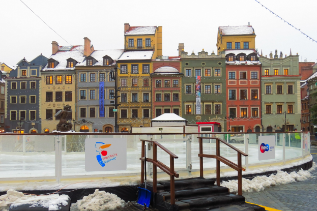 Eisbahn in einem Stadtplatz umgeben von Gebäuden, Treppen, Geländern, Informationsschildern, schneebedeckter Erde, Laternenmasten, Bäumen und Himmel.