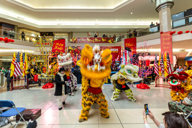 Ein lebhaftes chinesisches Neujahrsfest in einem Einkaufszentrum mit tanzenden Menschen in Kostümen und anderen, die auf Stühlen sitzen und mobile Telefone halten, umgeben von Flaggen, Bannern, Dekorationen und Deckenleuchten.