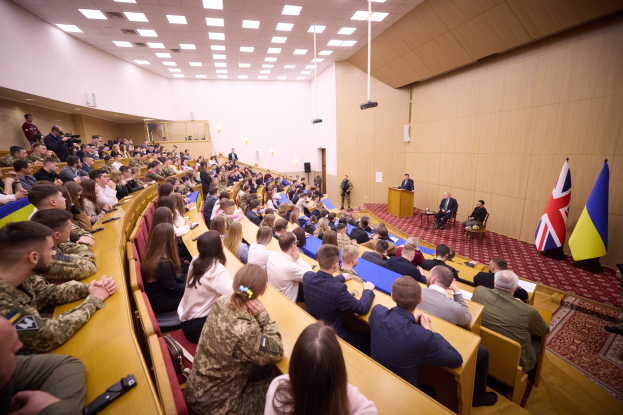 Große Gruppe von Menschen an Tischen in einem Raum mit Flaggen auf der rechten Seite, eine Wand im Hintergrund, Papiere und Gegenstände auf den Tischen, einigestehend in der Nähe eines Podiums mit einem Mikrofon, Deckenleuchten und ein Projektor.