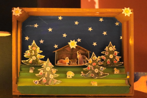 Eine hölzerne Krippe auf einem Tisch mit einer Lampe links und einer Wand im Hintergrund, dekoriert mit Weihnachtsbäumen, Tieren und einem Haus.