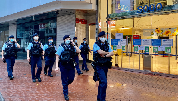 Eine Gruppe von Polizisten in Gesichtsmasken, die eine Straße vor einem gläsernen Gebäude entlanggehen, mit einem Geschäft auf der rechten Seite, das Plakate auf seinen Glaswänden hat.