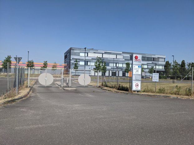 Großes industrielles Gebäude mit der Aufschrift "First Solar" und vielen Fenstern, umgeben von einem Metallzaun, umgeben von Bäumen, Pflanzen, Gras, Hinweistafeln, Straßenlaternen und einem Weg unter einem bewölkten Himmel.