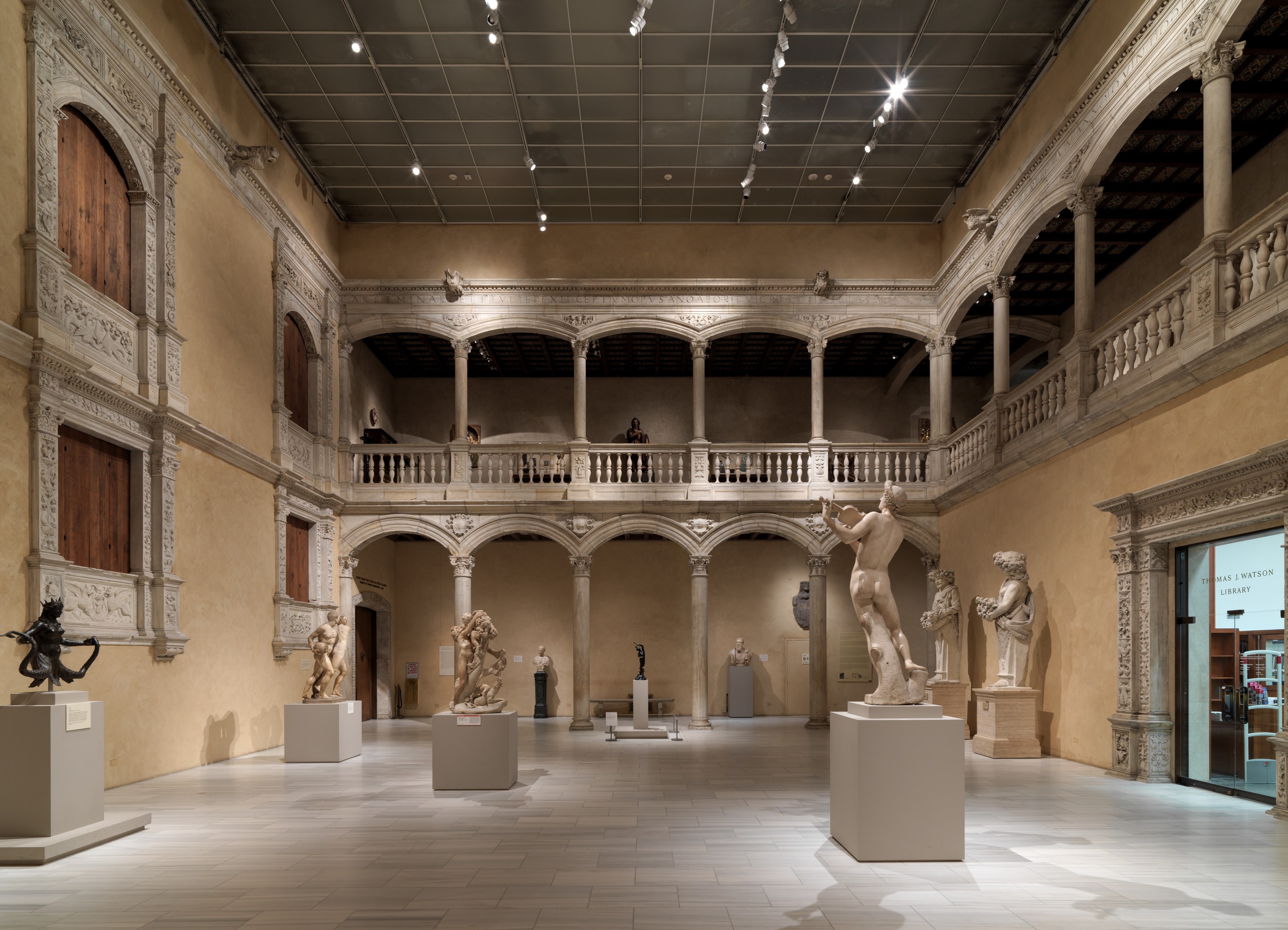Ein geräumiger Museumsgalerie mit zahlreichen Statuen und Skulpturen auf Sockeln, mit architektonischen Elementen wie Wänden, Säulen, Geländern und Deckenleuchten sowie einigen Besuchern.