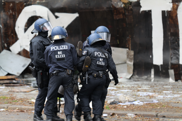 Polizeibeamte in schwarzen Uniformen und blauen Helmen stehen vor einem mit Graffiti beschmierten Gebäude während einer Unruhen, mit verstreuten Papieren und Gegenständen auf dem Boden.