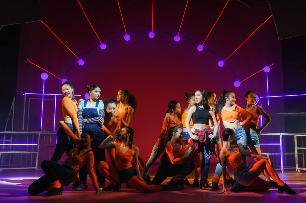 Eine Gruppe junger Tänzer in bunten Kostümen performs auf einer Bühne mit Geländern, beleuchtet von bunten Lichtern, mit einem Publikum im Hintergrund, das jubelt.