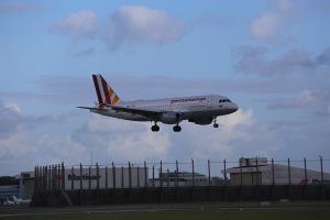 Germanwings Airbus A320-200 beim Start von Frankfurt Airport mit Gras darunter, einem Zaun im Vordergrund, Gebäuden und Bäumen im Hintergrund und einer bewölkten Himmel darüber.