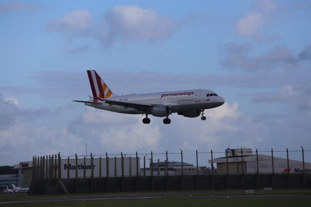Germanwings Airbus A320-200 beim Start von Frankfurt Airport mit Gras darunter, einem Zaun im Vordergrund, Gebäuden und Bäumen im Hintergrund und einer bewölkten Himmel darüber.