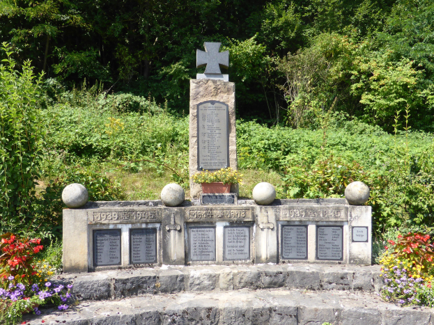 Denkmalstein mit Kreuz, beschriftet mit Wörtern und Zahlen, inmitten eines Gartens umgeben von Pflanzen und Bäumen, wahrscheinlich ein Weltkriegsgrab markierend.