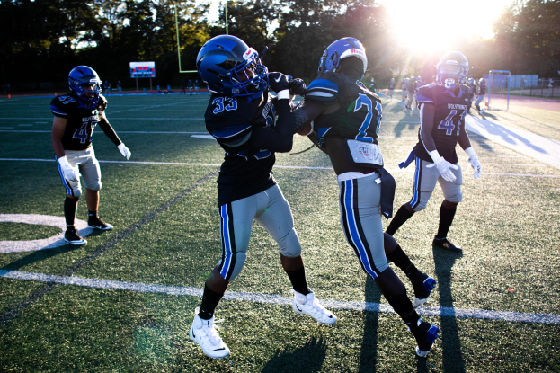 Gruppe junger Männer spielt Fußball auf einem Feld umgeben von Bäumen unter einem klaren blauen Himmel, trägt Helme und Schuhe.