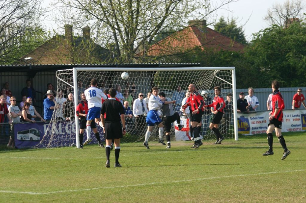 Fußballspieler spielen auf einem Feld mit einem Tor und Zuschauern hinter ihm, mit Bäumen und Häusern im Hintergrund.