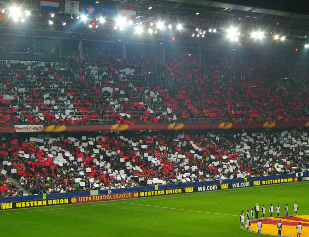 Ein Fußballspiel wird in einem großen, hell erleuchteten Stadion mit Zuschauern auf den Tribünen und Menschen auf dem Feld im Vordergrund gespielt.