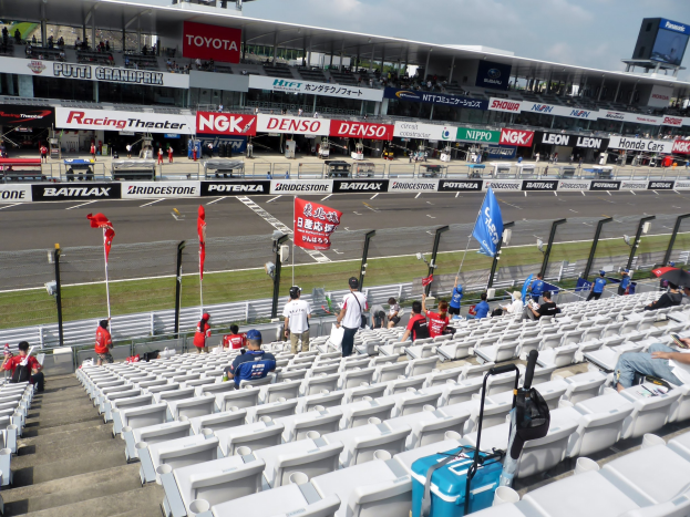 Eine Gruppe von Menschen sitzt auf Tribünen bei einer Rennstrecke mit Fahnen, Schildern, Geländern und Stufen, mit einem bewölkten Himmel über einem Stadion im Hintergrund.