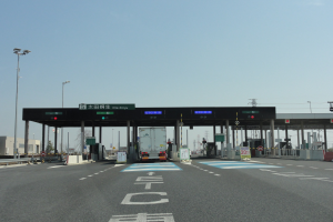 Mautstation mit einem vorfahrenden Lastwagen auf einer Straße mit weißen und blauen Markierungen, Laternen, Schildern und anderen Gegenständen, mit Gebäuden, Bäumen und einem klaren blauen Himmel im Hintergrund.