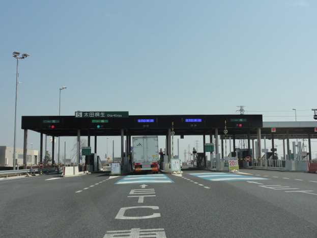 Mautstation mit einem vorfahrenden Lastwagen auf einer Straße mit weißen und blauen Markierungen, Laternen, Schildern und anderen Gegenständen, mit Gebäuden, Bäumen und einem klaren blauen Himmel im Hintergrund.