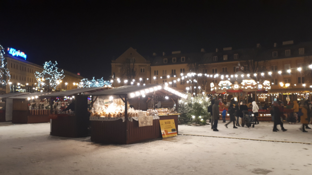 Ein nächtlicher Weihnachtsmarkt auf einer verschneiten Straße mit Menschen, beleuchteten Buden, Pflanzen, Bäumen, Gebäuden und Schildern unter einem bewölkten Himmel.