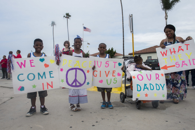 Eine Gruppe von Kindern hält Schilder in der Hand und steht vor einem Gebäude, während eine Frau einen Kinderwagen schiebt, umgeben von Bäumen, Pfählen, einer Flagge, einem Haus, Pflanzen und einem bewölkten Himmel; auf einem Schild steht 'Wir kommen in Frieden, gebt uns unseren Daddy'.