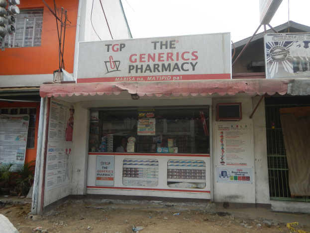 Eine Apotheke namens "Die Generics Apotheke" umgeben von Gebäuden, Strommasten, Werbetafeln, Hauspflanzen, Kraftfahrzeugen auf der Straße, unter einem klaren blauen Himmel.