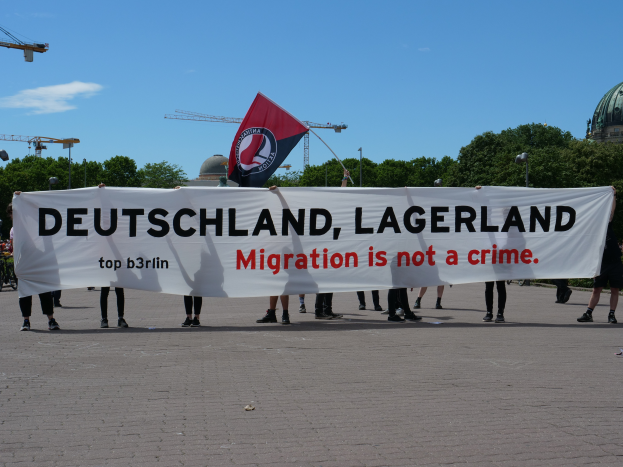 Eine Gruppe von Menschen mit einem Transparent, auf dem "Deutschland, Lagerland Migration ist kein Verbrechen" steht und einer Flagge, im Hintergrund Straßenlaternen, Bäume, Kräne, ein Gebäude und ein bewölkter Himmel.