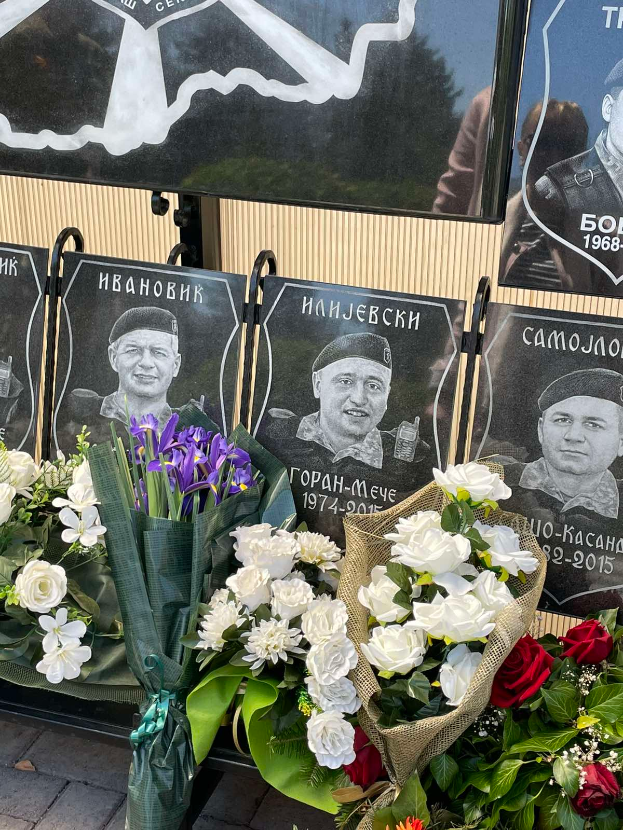 Ein Strauss bunter Blumen liegt auf einem Gehweg neben einem Schild, mit mehreren Gedenktafeln im Hintergrund.