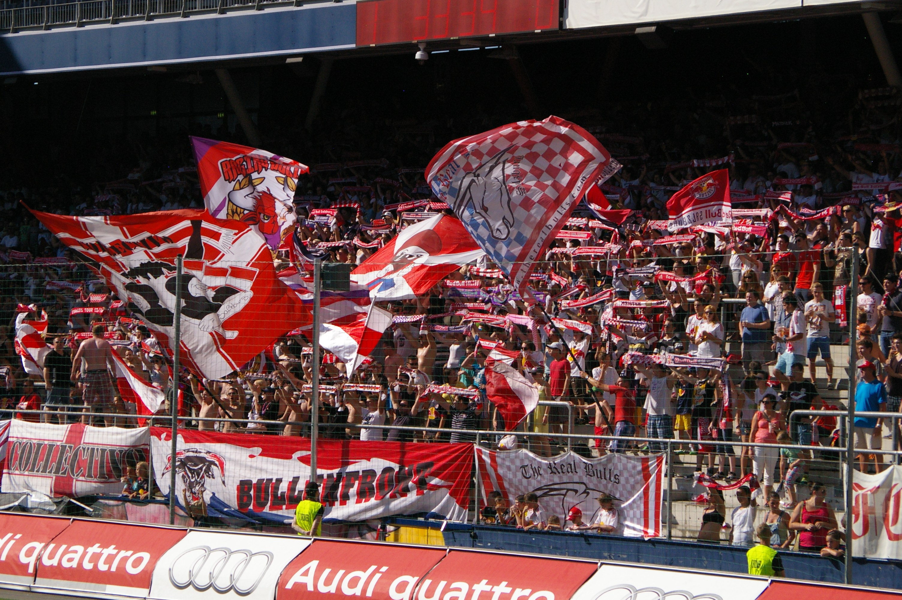 Große Menschenmenge in einem Stadion mit Fahnen, Bannern, einem Metallzaun, einem Schild, einem Anzeigebildschirm und Deckenleuchten.