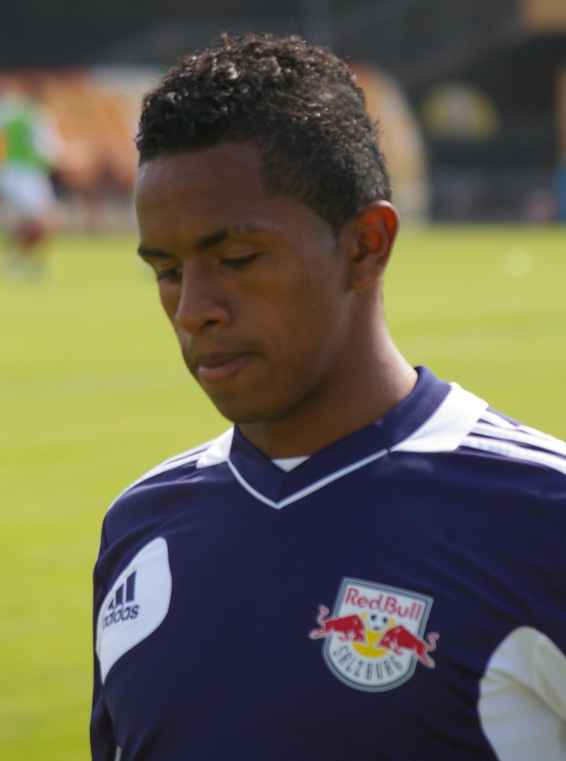 Ein junger Mann in einem blauen T-Shirt steht auf einem Fußballfeld, er scheint ein Flügelspieler mit kurzen dunklen Haaren und einem entschlossenen Gesichtsausdruck zu sein, vor einem unscharfen Hintergrund.