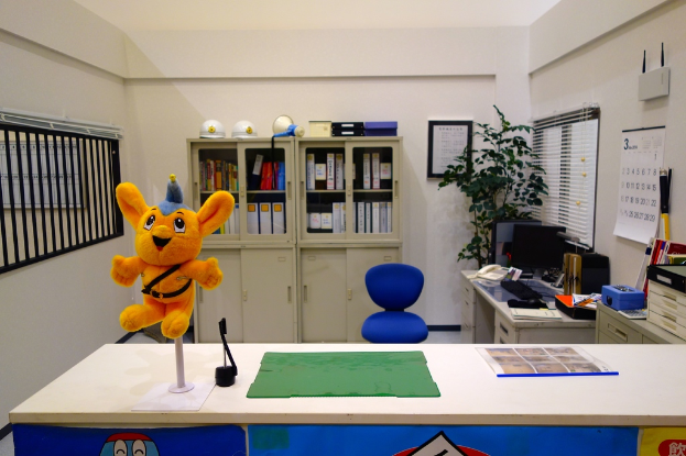 Ein Raum mit einem Tisch, auf dem eine Grafik und ein Teddybär auf einem Stab liegen, ein blauer Rollstuhl, ein Kalender an der Wand, ein Fenster, ein Topf mit einer Pflanze in der Ecke, ein Schrank mit einem Helm und einem Mikrofonsprecher, ein WLAN-Router und ein Desktop-Computer.