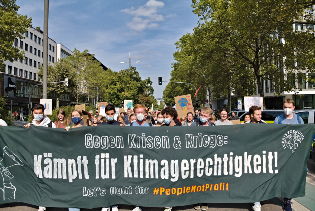 Eine Gruppe maskierter Individuals mit einem Banner, auf dem "Lass uns für die Menschen kämpfen, nicht für den Profit" steht, steht vor einer belebten Straßenszene mit Fahrzeugen, Bäumen, Gebäuden, Laternenmasten und Verkehrszeichen unter einem bewölkten Himmel.