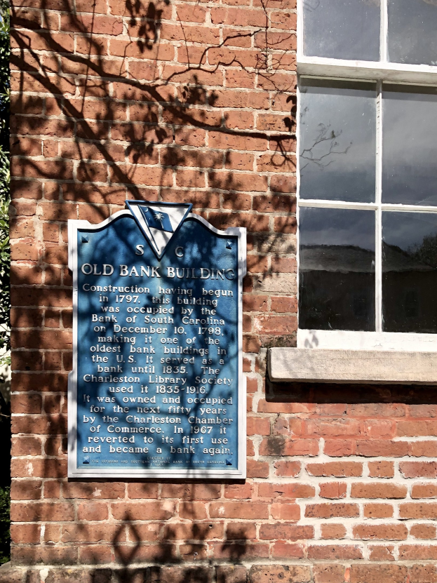 Altes Bankgebäude mit einer blauen Plakette an der Seite, ein Fenster auf der rechten Seite und ein Baum auf der linken Seite; die Plakette enthält Text.
