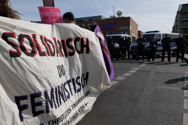 Eine Gruppe von Menschen marschiert auf einer Straße und hält ein "Solidarität und Feminismus"-Schild, mit parkenden Fahrzeugen und Gebäuden im Hintergrund unter einem klaren blauen Himmel.
