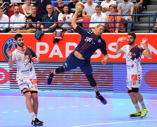 Eine Gruppe von Männern, die Handball auf einem Platz spielen, mit einem Ball in der Mitte, Zuschauern im Hintergrund und verstreuten Kleidungsstücken auf dem Platz, mit einem Banner, auf dem "Futsal-Weltmeisterschaft 2015 - Paris Saint-Germain vs Olympique Lyon" steht.