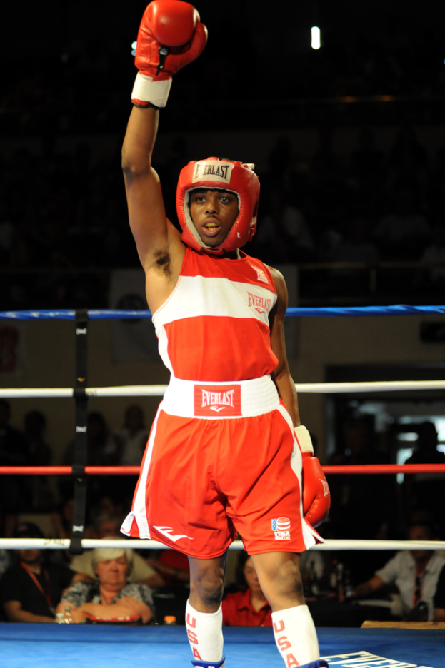 Ein Mann in Boxerbekleidung, einschließlich Helm und Handschuhen, steht in einem Boxring mit Zuschauern im Hintergrund.