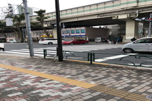 Stadtstraße mit Autos, Fußgängern an einer Ampel, Verkehrszeichen, einer Brücke, Bäumen, Gebäuden und einem klaren blauen Himmel.
